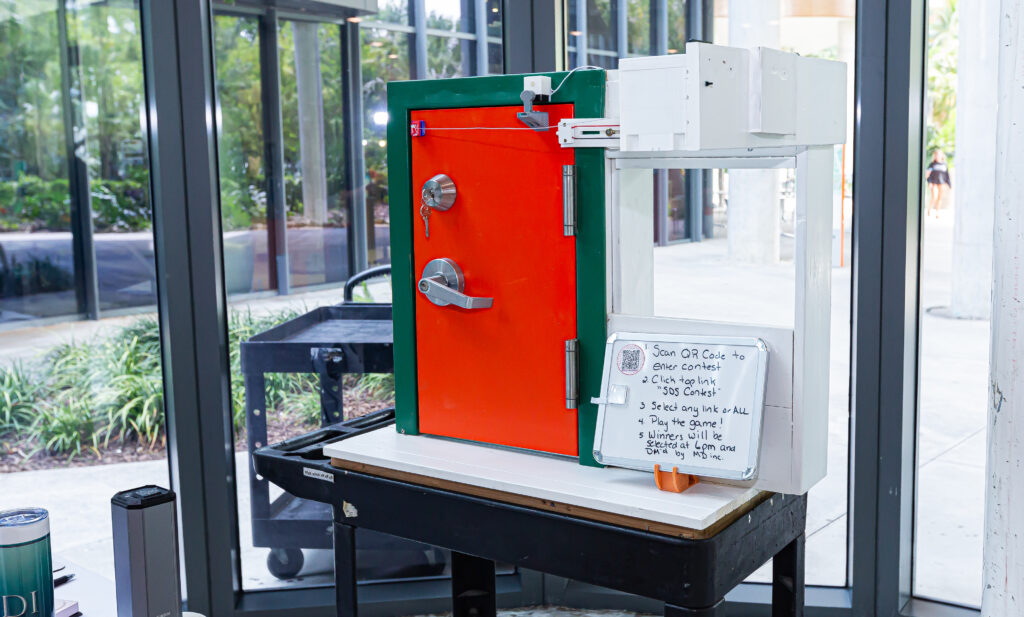 A prototype of the JERI Door Operating System, featuring a bold orange and green door with locking mechanisms, set on a display table. A small whiteboard next to the system provides instructions for interacting with the device.