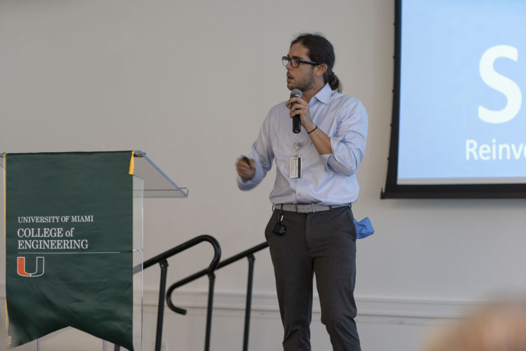 A presenter, identified as Joe, stands on stage at the 2023 Catalyzer Event at the University of Miami, speaking about Smart Door Solutions. He holds a microphone in one hand and gestures with the other, with a presentation screen in the background and a University of Miami College of Engineering banner on a nearby podium