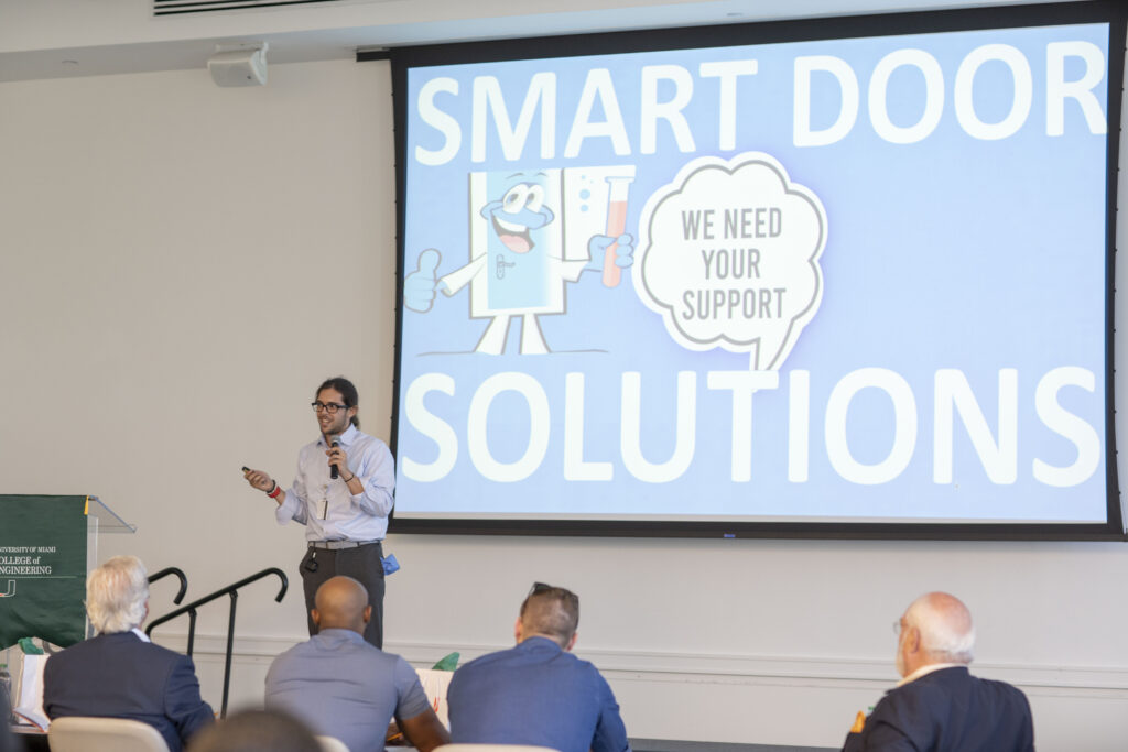 A presenter, identified as Joe, showcases the original logo of Smart Door Solutions during their first-ever pitch competition at the 2023 Catalyzer Event. The presentation slide behind him displays the logo with the tagline, "We Need Your Support," and an animated door character holding a test tube.